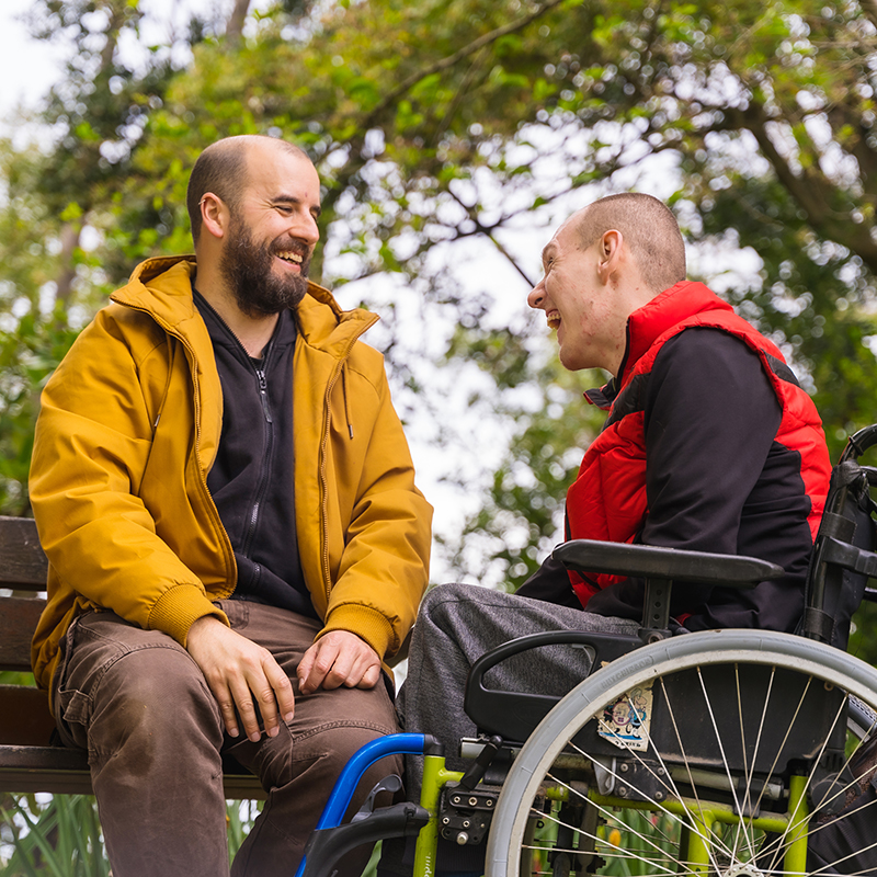 Porträt eines gelähmten jungen Mannes im Rollstuhl mit einem Freund auf einer Bank in einem öffentlichen Park der Stadt, die sich unterhalten und lachen.
